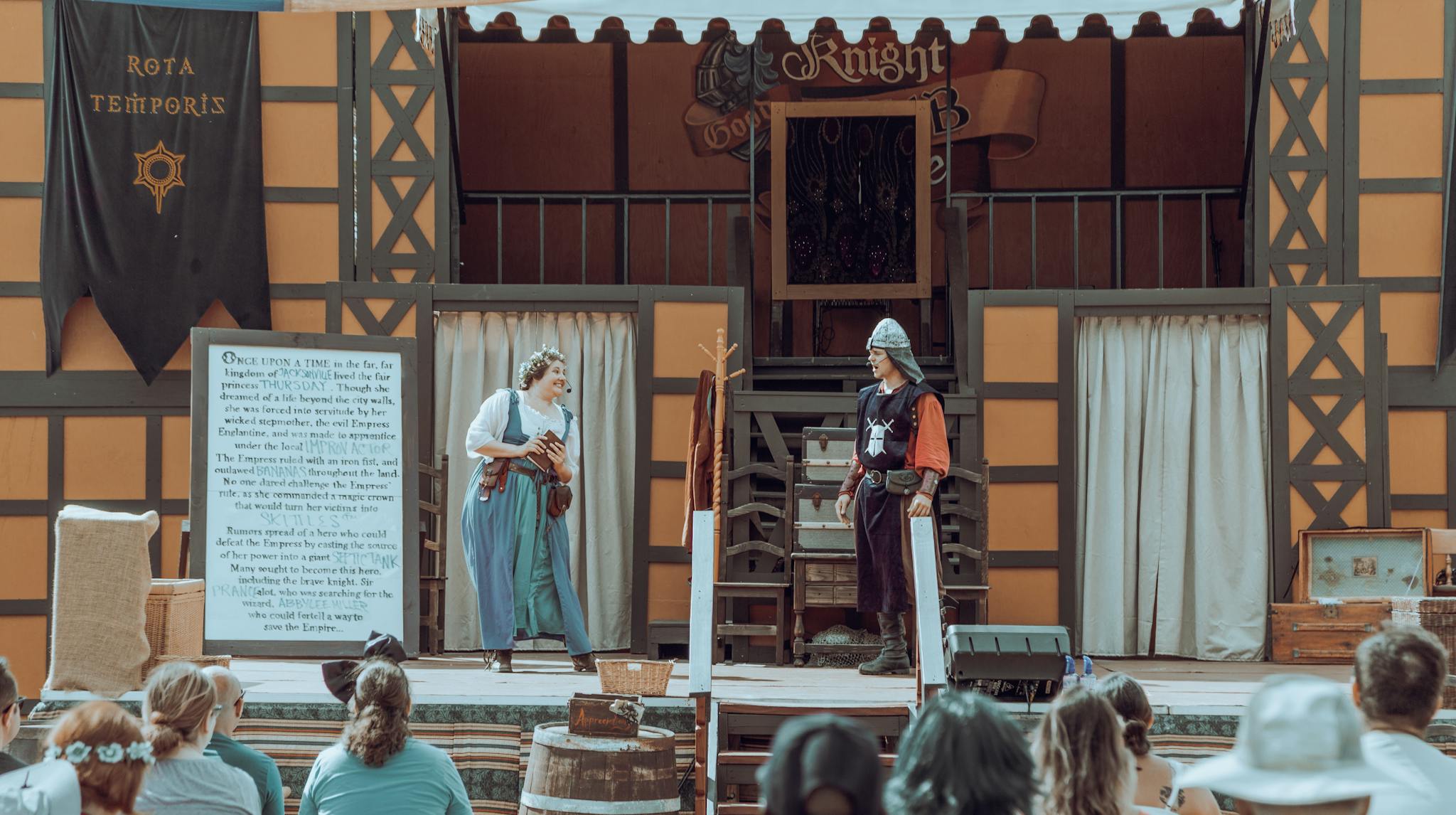 Actors in medieval costumes performing on an outdoor stage before an audience.